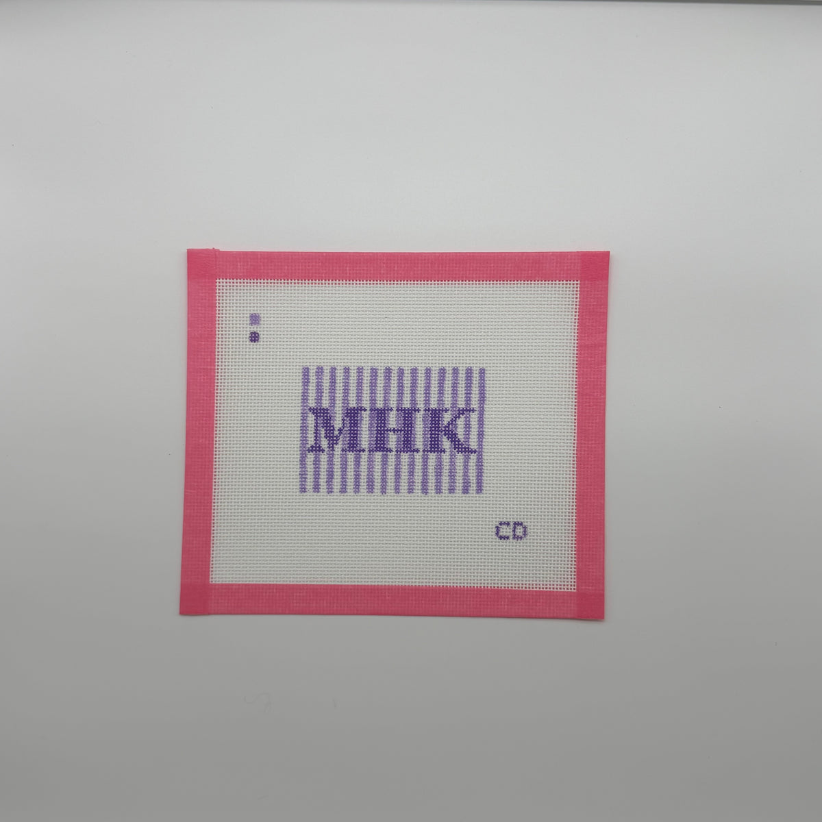 A square white cross-stitch canvas with a pink border, featuring purple embroidered letters 'MHK' inside vertical purple stripes, with a small purple dot and the letters 'CD' near the bottom right, shown on a light gray background.