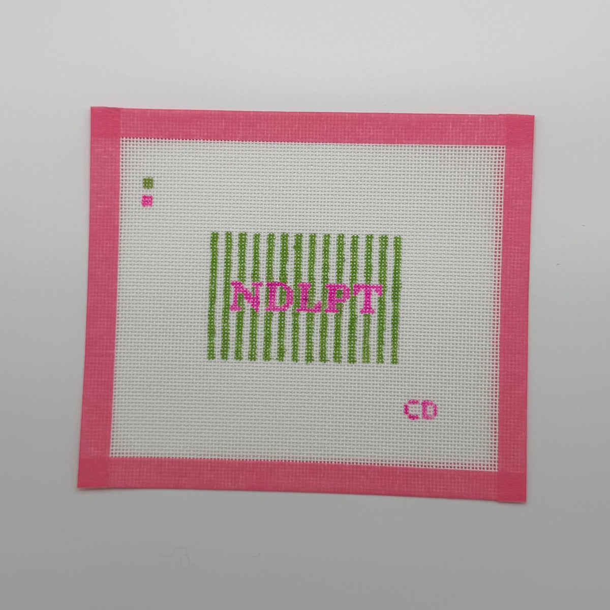 A square white embroidery canvas with a bright pink stitched border, featuring green vertical stripes, pink text reading 'NDEBT', and a small pink 'CD' in the lower right, shown on a plain light gray background.