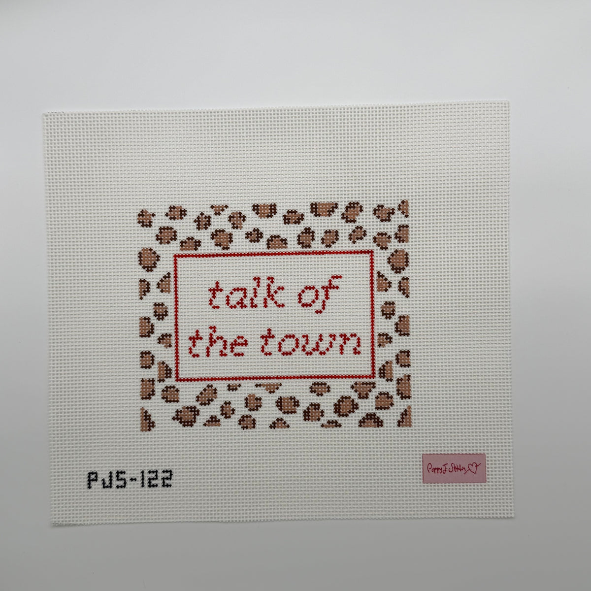 A square needlepoint canvas on white Aida fabric, featuring a leopard-print border in brown and beige with red stitched text reading 'talk of the town' inside a red rectangular frame. The canvas has a small pink brand label in the bottom right corner and the code 'PJ5-122' in black at the bottom left.